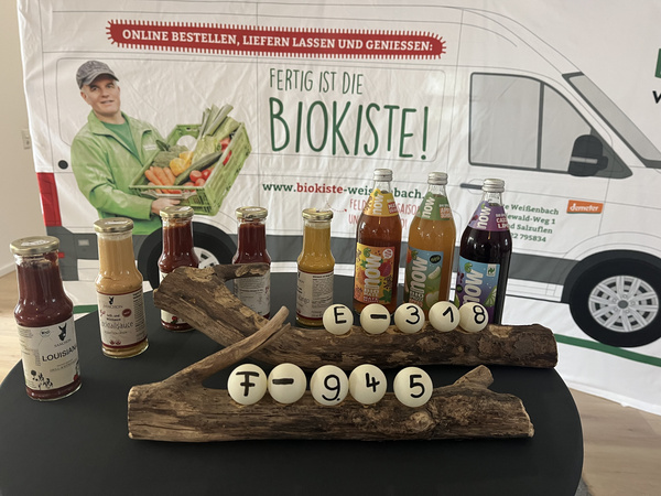 KI generiert: Das Bild zeigt einen dekorativen Ständer aus Holz mit darauf befestigten Kugeln, die Zahlen und Buchstaben tragen, sowie einen Geschenkkorb mit Lebensmitteln und Getränken im Hintergrund. Auf dem Ständer befindet sich auch ein Element mit Engelsflügeln.