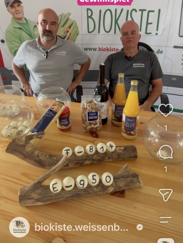 KI generiert: Das Bild zeigt einen dekorativen Ständer aus Holz mit darauf befestigten Kugeln, die Zahlen und Buchstaben tragen, sowie einen Geschenkkorb mit Lebensmitteln und Getränken im Hintergrund. Auf dem Ständer befindet sich auch ein Element mit Engelsflügeln.