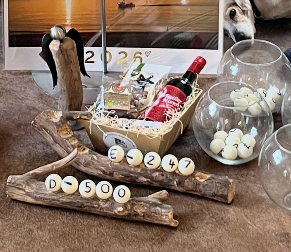 KI generiert: Das Bild zeigt einen dekorativen Ständer aus Holz mit darauf befestigten Kugeln, die Zahlen und Buchstaben tragen, sowie einen Geschenkkorb mit Lebensmitteln und Getränken im Hintergrund. Auf dem Ständer befindet sich auch ein Element mit Engelsflügeln.