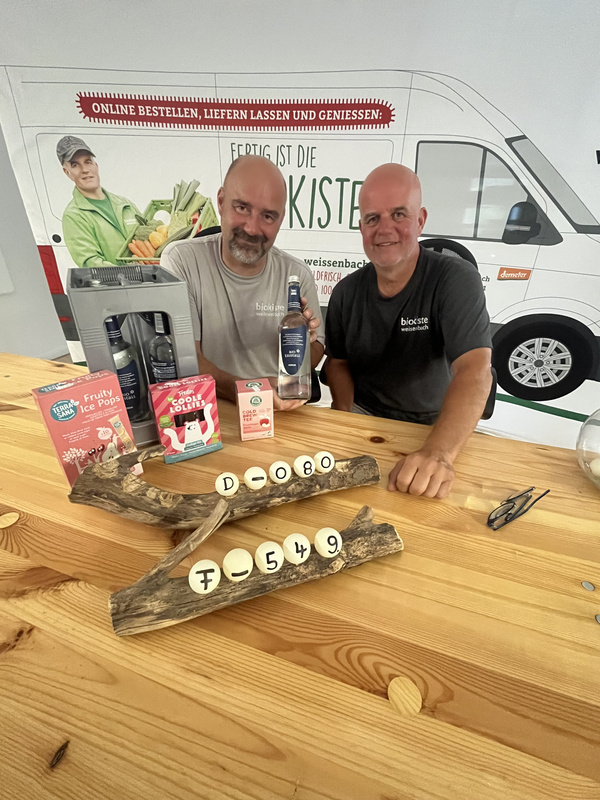 KI generiert: Das Bild zeigt einen dekorativen Ständer aus Holz mit darauf befestigten Kugeln, die Zahlen und Buchstaben tragen, sowie einen Geschenkkorb mit Lebensmitteln und Getränken im Hintergrund. Auf dem Ständer befindet sich auch ein Element mit Engelsflügeln.